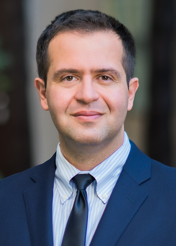 Headshot of Sina Houshmand, MD, man wearing a blue and white shirt, blue tie, blue jacket against a dark background