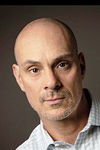 Professional headshot of a bald man with a trimmed beard, wearing a light checkered shirt and looking directly at the camera.