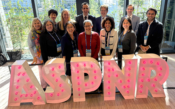 Group photo of UCSF faculty, alumni, and trainees standing indoors behind large illuminated ASPNR letters, smiling at the camera, with conference badges visible and a city view through floor to ceiling windows in the background.