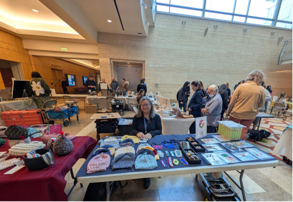 Cindy Cheng at a UCSF Artisan Guild By The Bay handmade fair. Woman smiling sitting behind a table of knitted hats and other handmade items.