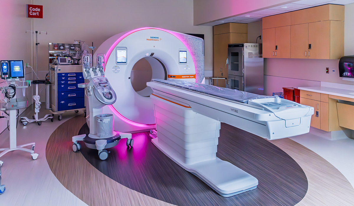 Photon-counting CT imaging suite with a Siemens scanner glowing with pink accent lighting, surrounded by medical equipment, cabinets, and a patient table in a clean clinical room