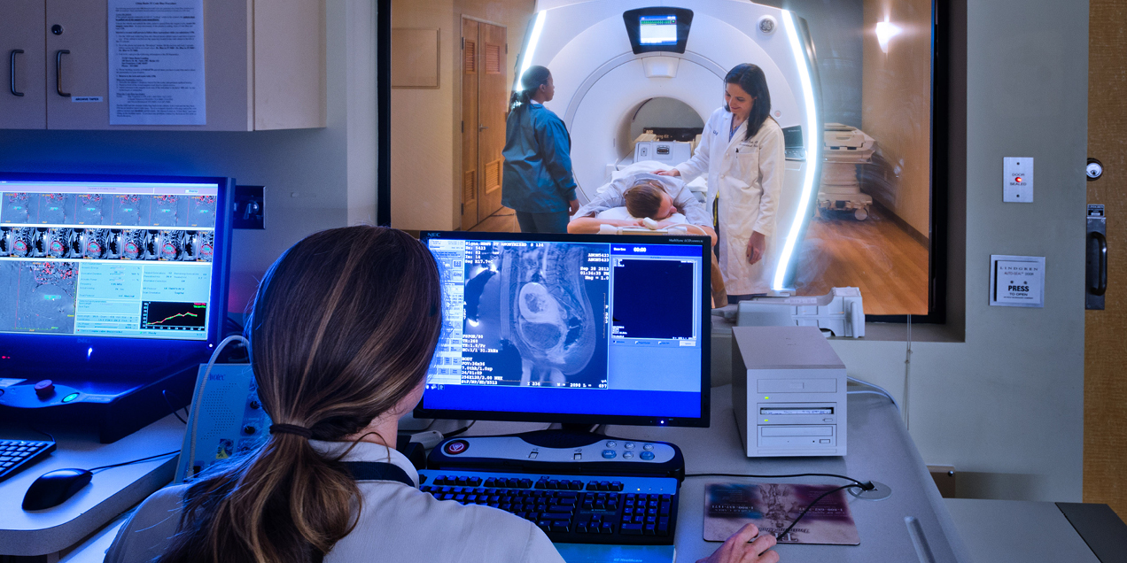 Clinical imaging control room with a technologist reviewing scan data on monitors while a patient is positioned in a scanner by clinicians in the adjacent imaging suite.