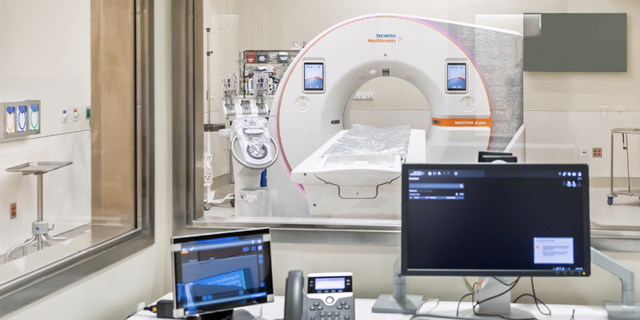 PET/CT scanner viewed through a control room window, with the imaging gantry centered in a clinical scanning suite and computer monitors and controls visible in the foreground.