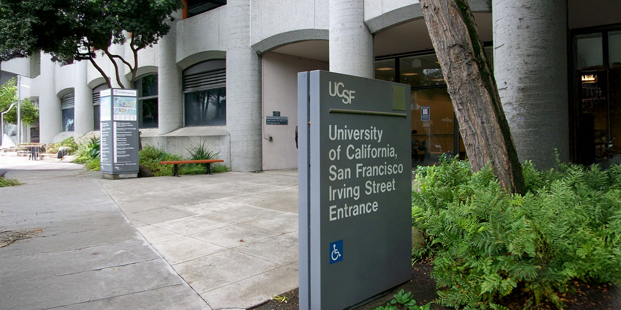 Exterior view of the UCSF Irving Street entrance, showing a campus walkway, concrete building columns, trees, and a UCSF wayfinding sign with accessibility symbol.