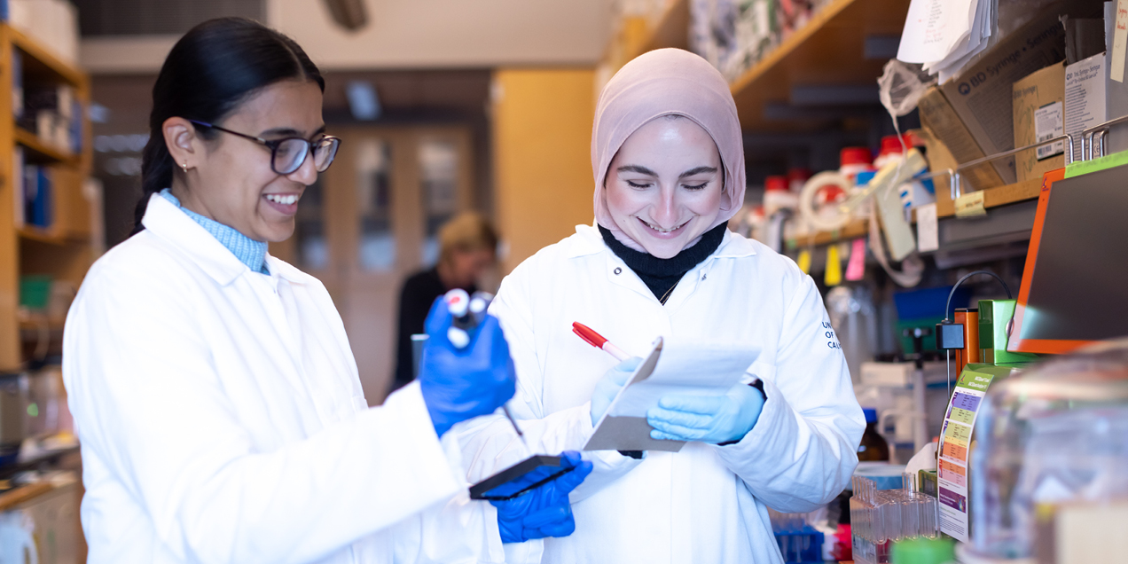 Two researchers in white lab coats smile while working together at a laboratory bench, reviewing notes and handling lab equipment.