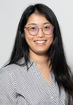 Headshot of a smiling person with long straight dark hair and round glasses, wearing a light gray patterned blouse and a delicate necklace against a light gray background.