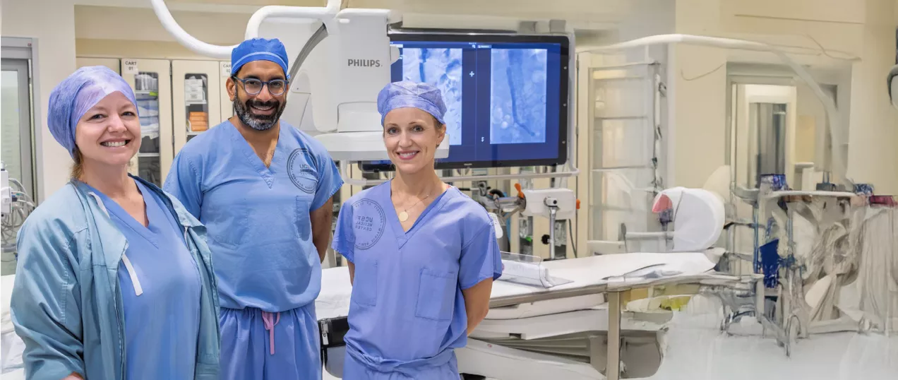 Three clinicians in blue surgical scrubs and caps stand smiling in a modern interventional radiology suite, with imaging monitors and medical equipment visible behind them.