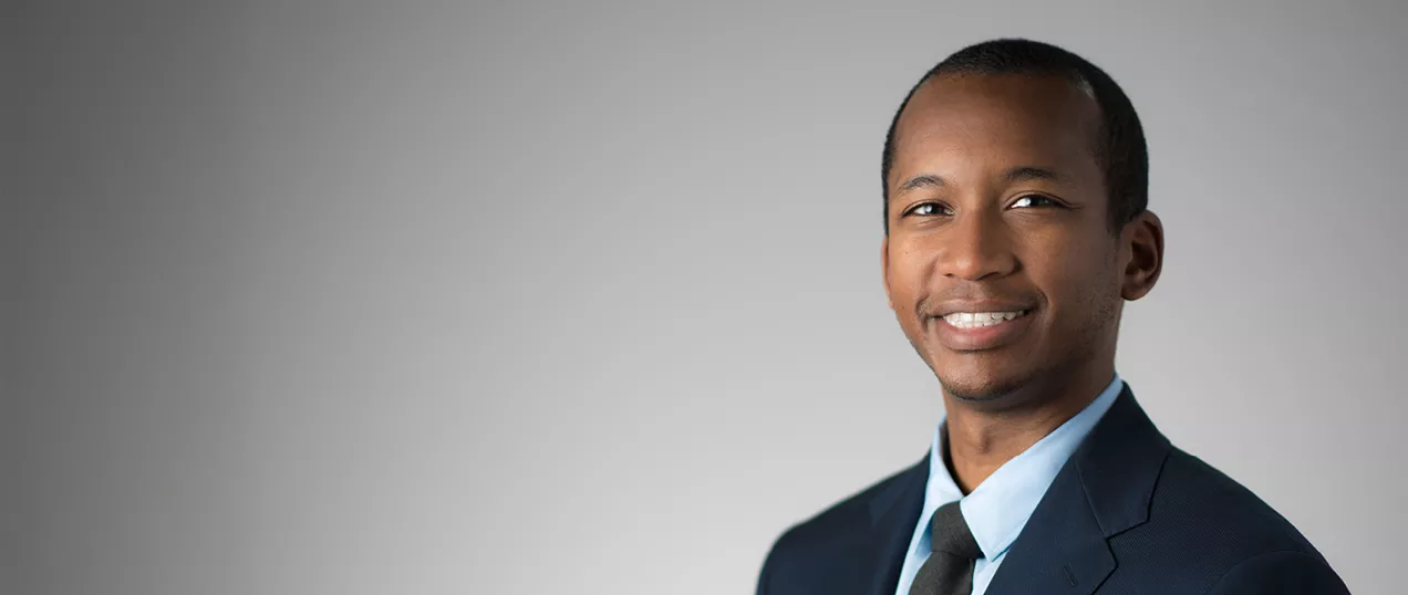 Professional headshot of a man smiling, wearing a dark suit, light blue shirt, and tie, against a neutral gray background