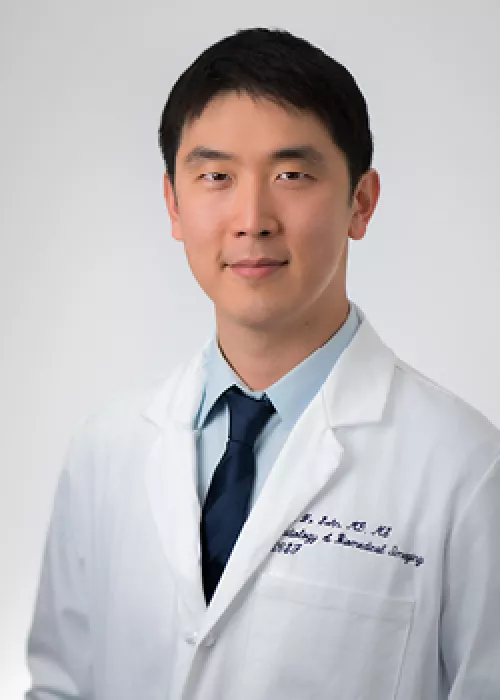 Jae Ho Sohn Portrait wearing a white lab coat and navy tie, standing against a light gray background.