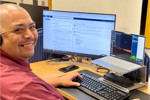 Smiling employee seated at a desk workstation, looking toward the camera while using a computer. A large monitor and laptop display work-related applications, with a keyboard, smartphone, and office equipment arranged on the desk in a professional office setting.