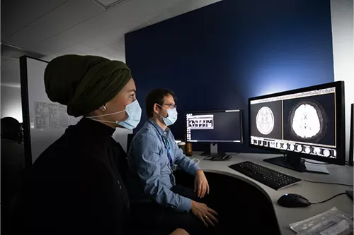 Two masked healthcare professionals seated in a radiology reading room, reviewing brain imaging studies on computer monitors. One person wears a dark green head covering and black clothing, while the other sits at a workstation displaying MRI scans. The dimly lit workspace is designed for medical image interpretation.