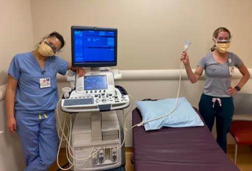 Two masked healthcare staff stand beside an ultrasound machine in an exam room, with one holding an ultrasound transducer next to an examination table.