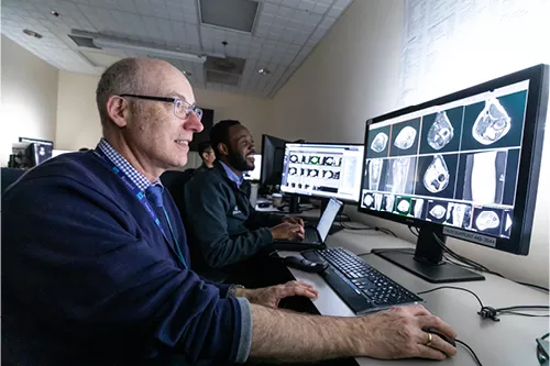 Alt text:  Two radiology professionals seated at computer workstations in a medical imaging reading room, reviewing diagnostic scans displayed on large monitors. The foreground monitor shows multiple cross-sectional medical images, while additional imaging studies are visible on nearby screens. The clinicians are engaged in image interpretation and collaborative review in a clinical workspace.