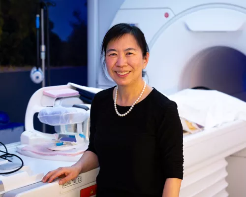 Professional portrait of a smiling woman wearing a black top and pearl necklace, standing beside an MRI scanner in a medical imaging suite.