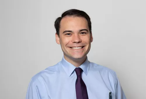 Professional headshot of a smiling man wearing a light blue dress shirt and dark tie, standing against a plain light gray background.