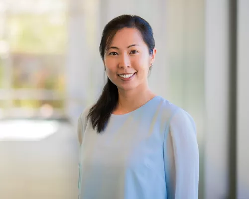 Professional headshot of a smiling woman wearing a light blue blouse, standing indoors against a softly blurred background with natural light.