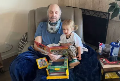 A man wearing a cervical neck brace reads a children's book with a young child while seated in a recliner, with toys and books nearby during his recovery at home.
