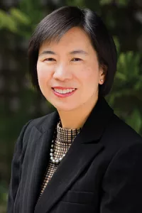 Professional outdoor headshot of Bonnie Joe wearing a black blazer and patterned blouse. She is smiling at the camera, with a softly blurred background of greenery.