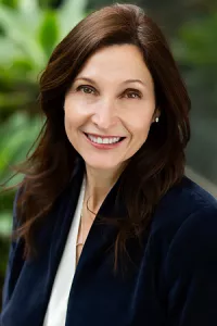 Professional outdoor headshot of Christine Glastonbury wearing a navy blazer over a light-colored blouse. She is smiling at the camera, with a softly blurred background of greenery.