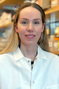Professional headshot of a woman with long blonde hair wearing a white laboratory coat. She faces the camera with a slight smile in a research or laboratory setting, with shelves and equipment softly blurred in the background.