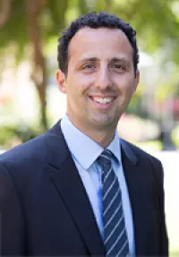 Portrait of a smiling person with short curly dark hair wearing a dark suit, light blue shirt, and blue striped tie. They are facing the camera outdoors with a softly blurred background of trees and greenery.