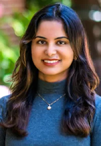 Professional headshot of a person with shoulder-length dark brown hair, smiling at the camera while wearing a navy blue mock-neck top and a delicate gold necklace with a small pendant. The portrait is set against a softly blurred outdoor background with green foliage.