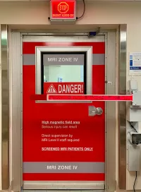 Red MRI safety door marking the entrance to MRI Zone IV, the restricted area containing the MRI scanner. Multiple warning signs indicate the presence of a strong magnetic field and caution that serious injury can result from unauthorized entry. A barrier across the door reads “Danger! Strong Magnet. Do Not Enter,” and a lighted sign above the doorway states “Stop. Magnet Always On.” The door is designated for screened MRI patients only and requires supervision by MRI-trained staff.