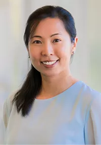 Head-and-shoulders portrait of a smiling woman with dark hair pulled back, wearing a light blue top, against a softly blurred outdoor background with greenery.