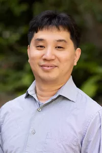 Professional outdoor headshot of Kang Wang wearing a light gray button-down shirt. He is smiling at the camera, with a softly blurred background of greenery.