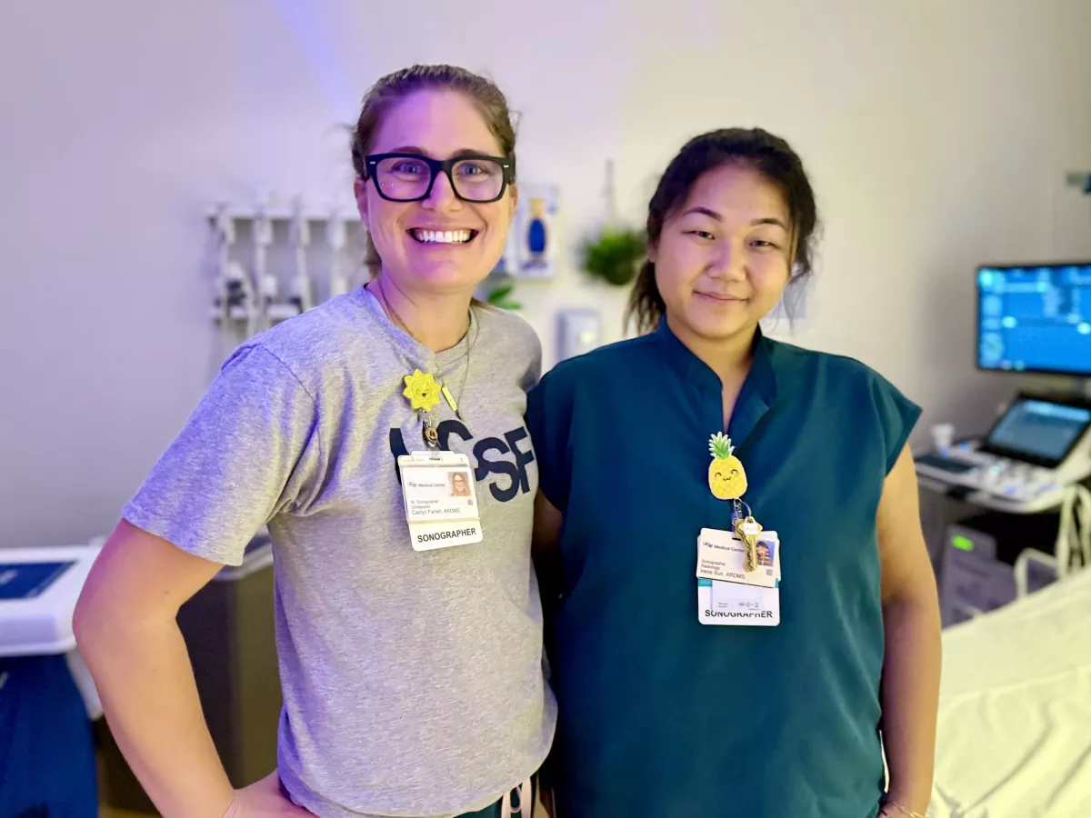 Two UCSF sonographers smiling together in a dimly lit exam room. One wears glasses and a UCSF t-shirt, and the other wears teal scrubs; both have ID badges with cheerful badge decorations. An ultrasound machine sits in the background.