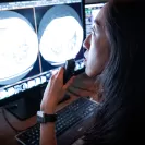 adiologist examining CT scan images on dual computer monitors in a dark reading room. She holds a microphone for dictation while closely analyzing the scans, with the glow of the screens illuminating her focused expression.