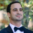 Portrait of a man wearing a black tuxedo and bow tie, outdoors with a blurred natural background.