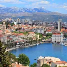 Panoramic view of Split harbor with red roofed historic buildings, waterfront promenade, boats, and mountains in the background.