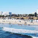 Wide view of a sunny beach with people swimming and walking along the shoreline, ocean waves in the foreground, and a row of palm trees and tall buildings lining the coast in the background.