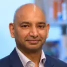 Portrait of a bald man wearing a suit jacket and collared shirt, looking at the camera with a neutral expression against a softly blurred indoor background.