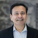 Headshot of a man smiling, wearing a dark blazer over a light collared shirt, set against a softly blurred gray background.