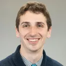 Headshot of a smiling man with short brown hair, wearing a collared shirt, tie, and dark sweater, against a neutral gray background.