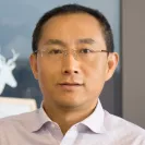 Professional headshot of a man wearing glasses and a light-colored button-up shirt, facing the camera with a neutral expression. The background is softly blurred with framed artwork visible behind him.