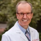Professional headshot of a smiling man wearing glasses, a white medical coat, a light blue dress shirt, and a patterned tie. He is facing the camera against a softly blurred outdoor background with greenery. The portrait has a polished and approachable appearance suitable for a physician, faculty, or leadership profile.