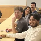Four people seated in a classroom or conference room smile toward the camera from tiered desks. In the foreground, a person in a white hoodie smiles broadly, while another person in a gray T-shirt looks back over their shoulder with their chin resting on one hand and a phone in the other. Two more people sit behind them, smiling. Laptops, phones, and office chairs are visible, suggesting a seminar or meeting setting.