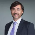Professional headshot of Riccardo Lattanzi wearing a dark suit and lavender tie, smiling against a gray background.