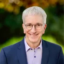 Head-and-shoulders portrait of an older man with short gray hair and round glasses, smiling at the camera. He wears a navy blazer over a light lavender shirt, with softly blurred green and golden foliage in the background.