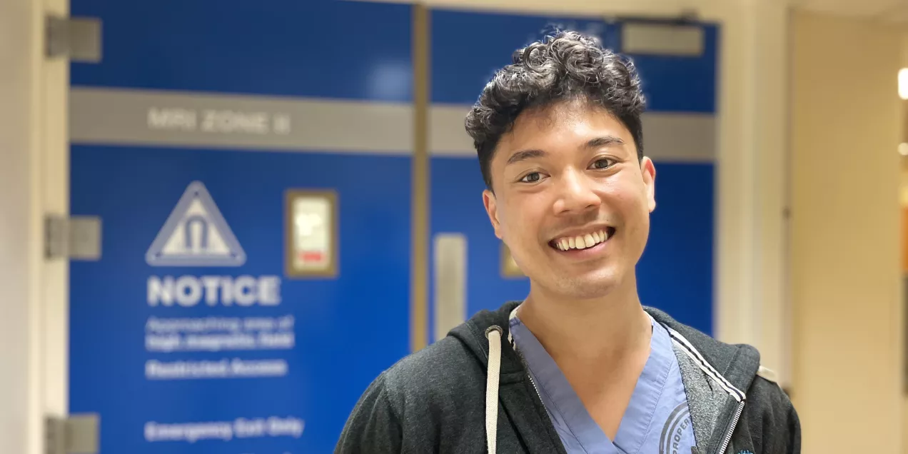 A medical professional in scrubs and a zip-up hoodie smiling in a hospital hallway, standing in front of blue MRI Zone II doors with safety signage.
