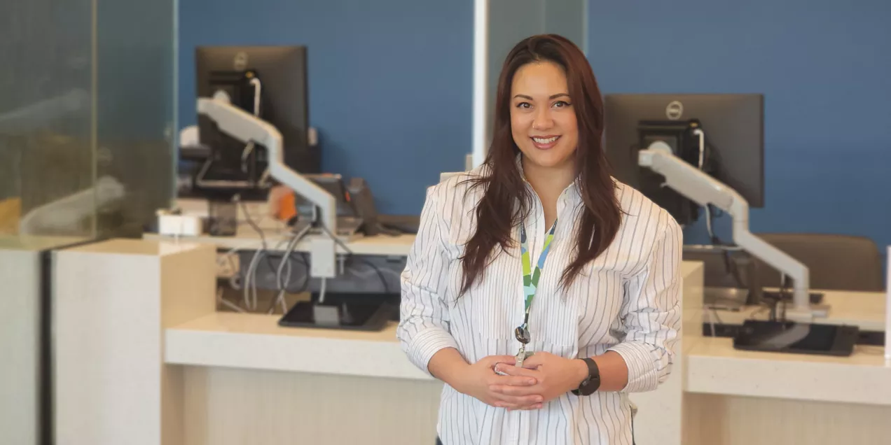 A staff member standing in the lobby of a medical outpatient center, smiling in front of a counter with computer workstations and a blue wall displaying the facility’s name.