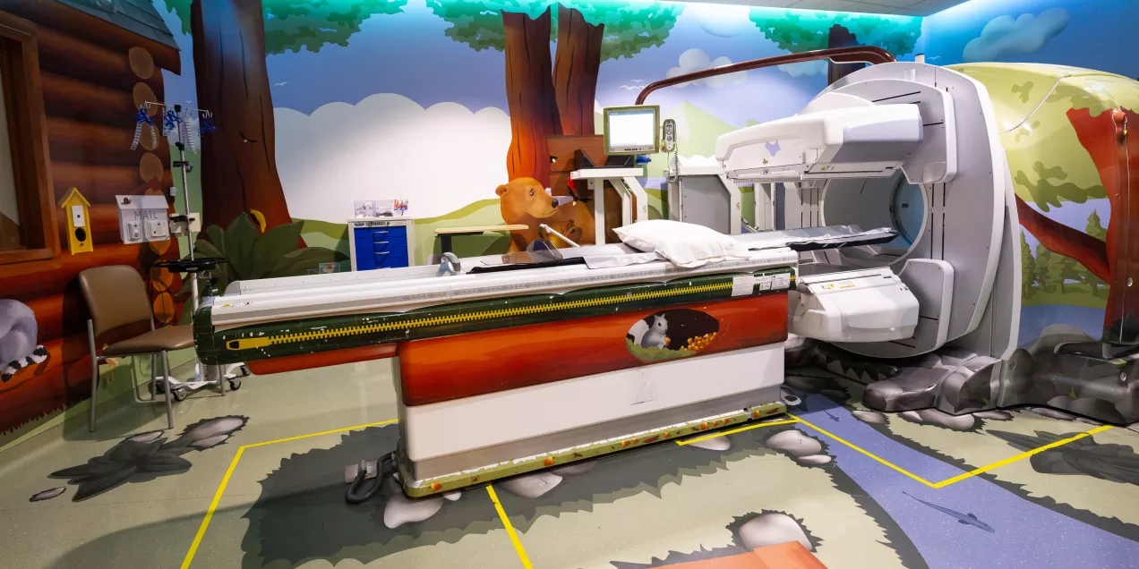 A pediatric imaging room decorated with a forest theme, featuring a large scanner surrounded by colorful murals of trees, animals, and nature. The exam table and medical equipment are integrated into the playful woodland design.