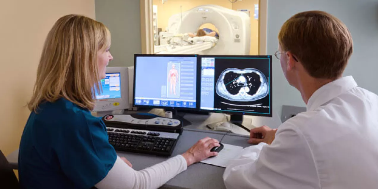 two clinicians in a control room reviewing CT scan images on dual monitors while a patient undergoes a scan in a nearby imaging suite visible through the window