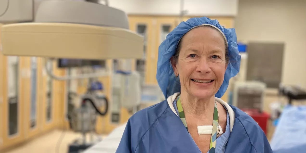 A medical professional in blue surgical scrubs and a blue bouffant cap smiling in a procedure room, with medical equipment and cabinets in the background.
