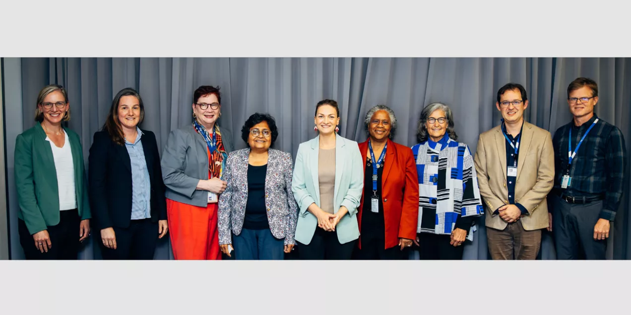 Group of nine professionals standing in a row in front of a gray curtain, smiling at the camera. They are dressed in business and professional attire, wearing a mix of jackets, blazers, and ID badges, representing a formal or academic gathering.