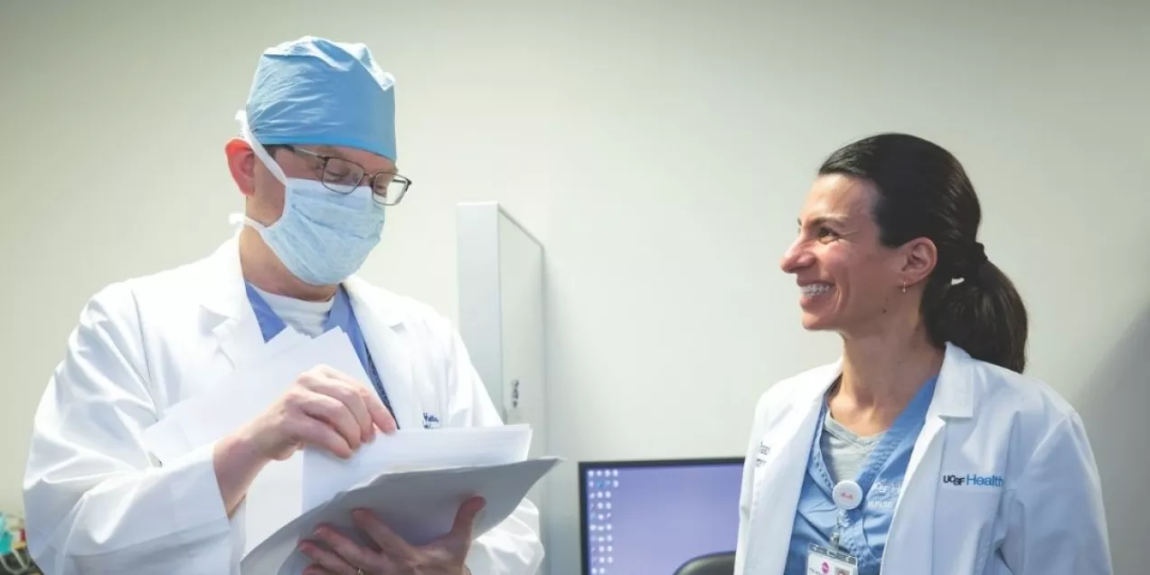 Two UCSF Health clinicians in scrubs and white coats talk in a clinical workspace, with one reviewing papers and the other smiling.