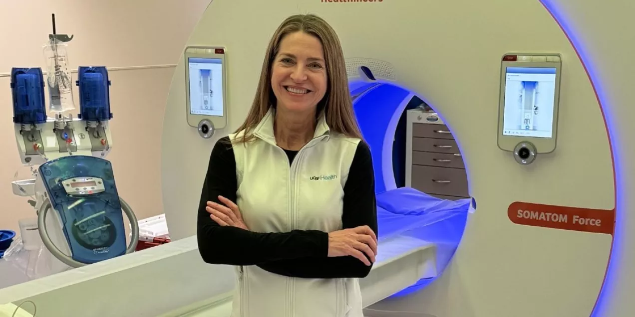 A woman stands in front of a Siemens Healthineers SOMATOM Force CT scanner with her arms crossed, smiling. She’s wearing clinical work pants, sneakers, and a white vest over a black long-sleeve shirt. Additional medical equipment is visible beside the scanner in a clean imaging room.